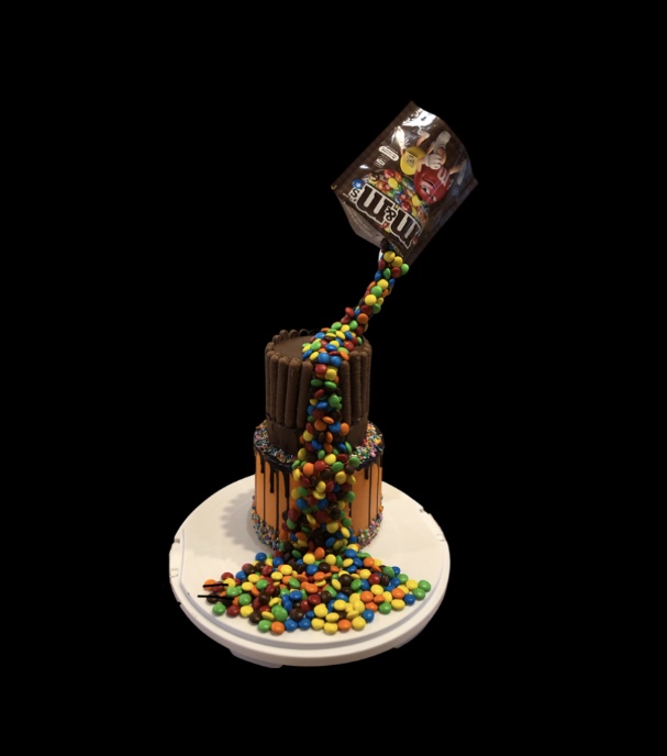 3D illusion waterfall cake with M&Ms pouring from the packet over a chocolate and orange two layer cake with chocolate fingers and sprinkles
